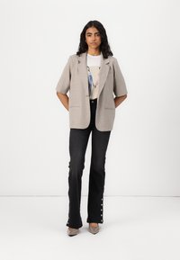 Světle šedý oversized blazer s krátkými rukávy, v kombinaci s bílým grafickým tričkem a černými zvonovými džíny s knoflíkovými detaily.
