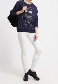 Felpa blu navy con grafiche dorate che riportano "LONDRA", "NEW YORK" e "PARIGI", abbinata a jeans skinny bianchi e sneakers nere.