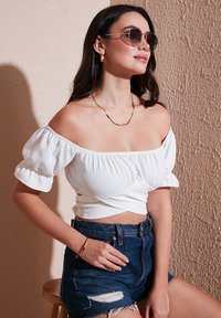 Hvid off-the-shoulder top med puffede ærmer, tekstureret stof, parret med mørke denim shorts og diskrete guldsmykker, på en neutral baggrund.