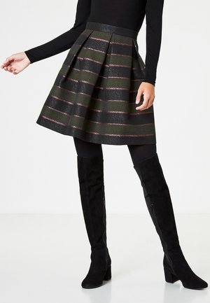 Femme portant un haut noir à manches longues, une jupe plissée noire avec des rayures horizontales roses, des collants noirs et des bottes noires à talons hautes jusqu'aux genoux.