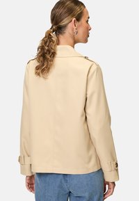 Beige lättviktsjacka med rak skärning, stor krage och knappaccenter på axlar och ärmslut. Slät textur, inga mönster.