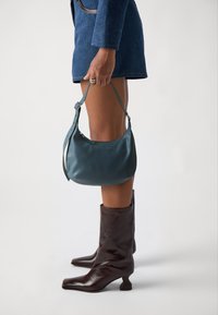Bolso de cuero color azul pálido con forma curvada y correa ajustable, sostenido por una persona que lleva una chaqueta azul y botas de tobillo marrones.