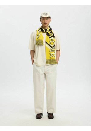 Mees seisab käed taskutes, kandes kreemikat särki, laia valget pükst, valget mütsi, kollase ja musta mustriga sall ning pruune kingi.