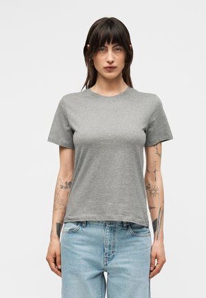 Naine tume juustega, kandes tavalist halli lühikeste varrukatega T-särki ja helesiniseid teksaseid, seistes valge tausta ees.