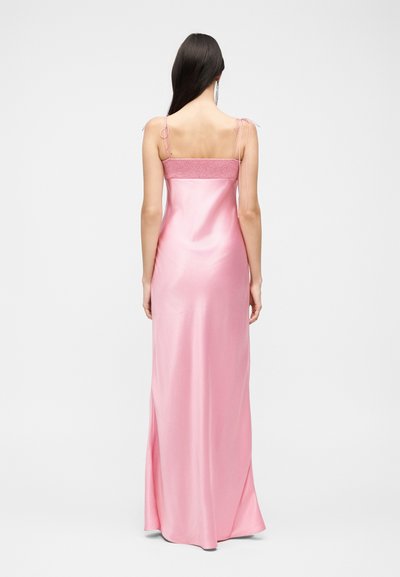 Γυναίκα με μακριά σκούρα μαλλιά που φοράει ένα μακρύ, γυαλιστερό ροζ σατέν φόρεμα με λεπτές τιράντες δεμένες σε φιόγκους, με την πλάτη στραμμένη.
