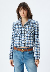 Skuren jacka med blå och vit rutig mönster, tillverkad av strukturtyg. Har två bröstfickor och knappar fram.