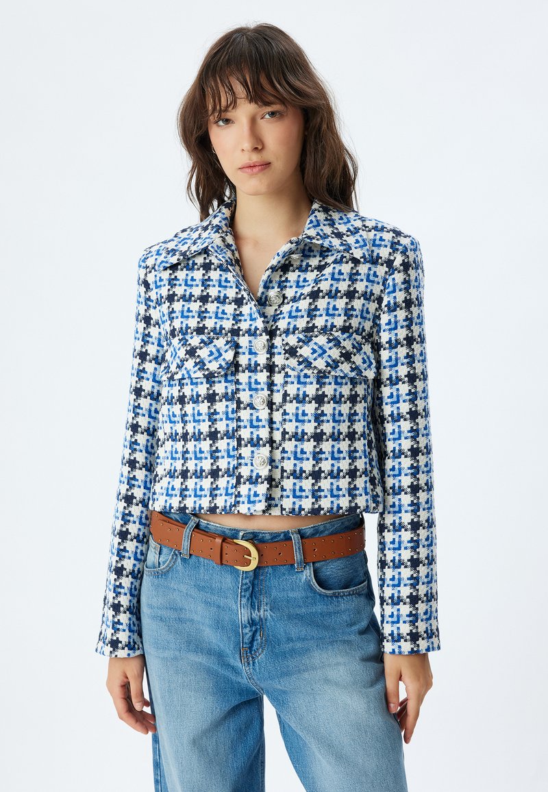 Skuren jacka med blå och vit rutig mönster, tillverkad av strukturtyg. Har två bröstfickor och knappar fram.