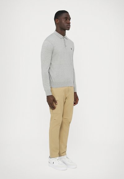 Szürke hosszú ujjú galléros póló, bézs színű cargo nadrág és fehér tornacipő. Simább anyagok és egyszerű kialakítás. Kis logó a pólón.