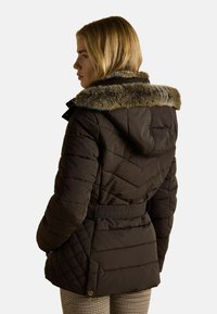Hnedá prešívaná bunda s kapucňou, so zatepleným kožúškom, opaskom v páse a textúrovanou tkaninou. Pohľad zozadu ukazuje štepovanie v diamantovom vzore.