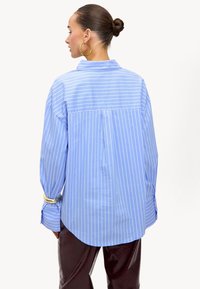 Lichtblauw, oversized shirt met witte verticale strepen, een achterplic en knoopmanchetten. De stof lijkt glad en licht van gewicht.