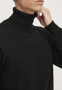 Schwarzer Rollkragenpullover mit strukturierten horizontalen Streifen, geripptem Kragen und Bündchen. Das Material wirkt weich und ist gestrickt.