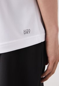 Vit andningsbar skjorta med mesh-textur, med "ULTRA DRY" i grått. Parat med svarta tygbyxor, fokus på skjortans fålldetalj.