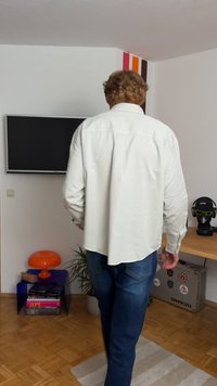 Camisa de manga longa bege claro com uma bainha curva e colarinho clássico, combinada com jeans azul-escuro. O fundo apresenta uma televisão e uma decoração colorida.
