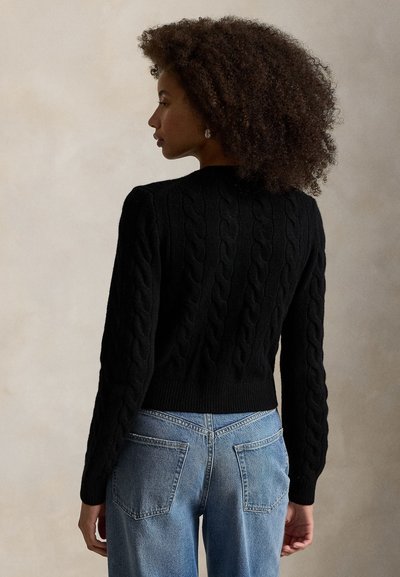 Kvinde med krøllet hår iført en sort kabelstrikket sweater og lyseblå jeans, der står med ryggen til mod en ensfarvet baggrund.