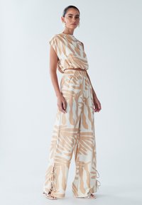 Lins auduma divdaļīgs komplekts ar bēša un baltas krāsas abstraktu rakstu. Platām kāju biksēm ar elastīgu jostasvietu un sānu auklām; īss tops ar asimetrisku dizainu.