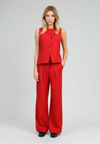 Femme se tenant contre un fond gris, portant un gilet rouge à boutons sans manches et un pantalon large rouge avec des chaussures noires.