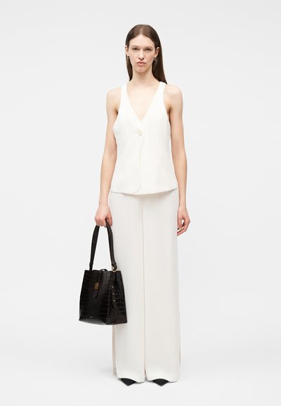 Γυναίκα με λευκό αμάνικο γιλέκο και λευκό παντελόνι φαρδύ, κρατώντας μια μαύρη τσάντα με υφή και δύο λαβές, στέκεται μπροστά σε μια απλή φόντο.