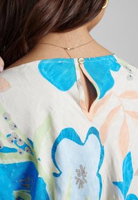 Dos d'une femme montrant une encolure à agrafe sur une blouse blanche avec un grand imprimé floral bleu et pêche, collier et boucle d'oreille en or visibles.