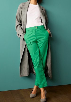 Grijze lange jas, wit T-shirt en felgroene broek. De broek is recht gesneden met zakken; de schoenen zijn lichtgrijs met een puntige neus.