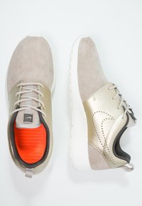 Women's sneakers featuring a beige suede upper, metallic gold accents, white rubber sole, and bright orange inner lining.