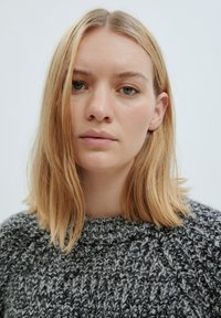 Grauer und schwarzer Strickpullover mit strukturiertem Muster. Rundhalsausschnitt und leicht lockere Passform. Das Modell hat schulterlange blonde Haare.