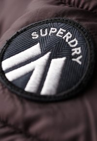 Une patch circulaire noire avec le texte "SUPERDRY" et des éléments graphiques en blanc en gras sur un fond en tissu sombre texturé.