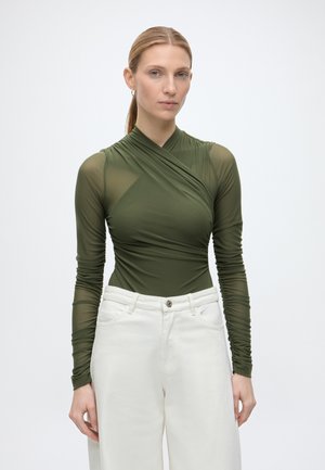 Femme à la peau claire et aux cheveux blonds porte un haut moulant vert olive à manches longues et un pantalon blanc ample, debout devant un fond uni.