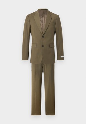 Άνδρας με σγουρά μαλλιά που φοράει ελαιί πράσινο κοστούμι, λευκό πουκάμισο, μπεζ γραβάτα και μαύρα επίσημα παπούτσια, στέκεται με το ένα χέρι στην τσέπη.