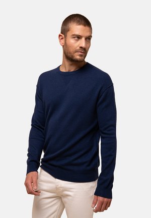 Pull bleu marine en tissu doux, design à col rond, manches longues et poignets côtelés avec une texture lisse. Porté par-dessus un pantalon clair.