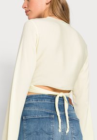 Top cropped amarelo claro com mangas longas e um laço nas costas, combinado com calças de ganga azuis com dois bolsos traseiros.
