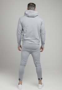 Hombre con tatuajes, vistiendo una sudadera ligera gris y pantalones jogger a juego, de pie dando la espalda contra un fondo gris claro liso.