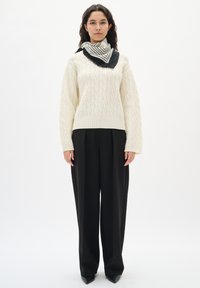 Kvinde iført en cremefarvet kabelstrikket sweater, sorte vide bukser, sorte spidse sko og et sort-hvidt stribet tørklæde.