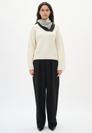 Frau, die einen cremefarbenen Kabelstrickpullover, schwarze weit geschnittene Hosen, schwarze spitze Schuhe und einen schwarz-weißen gestreiften Schal trägt.