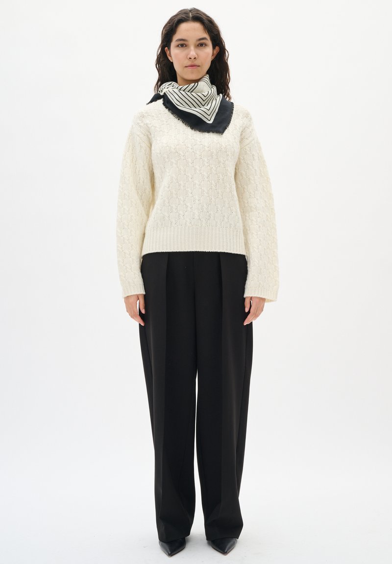 Mujer vestida con un suéter de punto en color crema, pantalones de pierna ancha negros, zapatos de punta negros y una bufanda a rayas en blanco y negro.