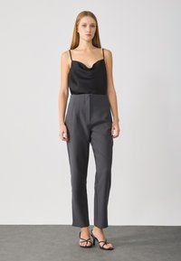 Schwarzes Satin-Camisole mit Cowl-Ausschnitt, kombiniert mit hochtaillierten, maßgeschneiderten grauen Hosen. Das Modell trägt schwarze Sandalen. Glatte Texturen überall.
