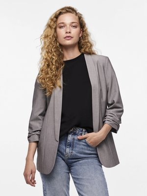 Donna con lunghi capelli biondi ricci, che indossa un blazer grigio, una camicia nera e jeans azzurri, posa con una mano in tasca su sfondo bianco.