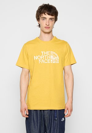 Giovane uomo che indossa una t-shirt gialla The North Face e pantaloni blu scuro, in piedi davanti a uno sfondo chiaro e uniforme.