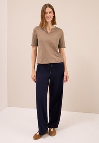 En smilende kvinde står iført en brun kortærmet top, marineblå brede bukser og brune sneakers foran en ensfarvet beige baggrund.