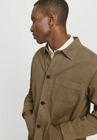 Chaqueta de lona marrón con diseño de cuello, que presenta botones en la parte delantera, un bolsillo en el pecho y dos bolsillos laterales. Tejido texturizado.