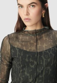 Giovane donna con occhi verdi che indossa una blusa trasparente a fantasia scura e grandi orecchini a cerchio dorati, guardando leggermente di lato.