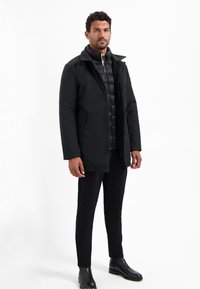 Zwarte pufferjack met een kraag, vooraan met drukknoopsluiting en een gladde buitenkant. Draagt over strakke zwarte broek en enkellaarsjes.