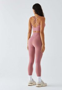 Różowy sportowy biustonosz z krzyżującym się tyłem, zestawiony z różowymi legginsami z wysokim stanem. Sneakersy z jasnymi akcentami, białe skarpetki do kostek. Gładka faktura.