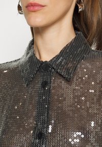 Camicia nera di paillettes con colletto classico, realizzata in tessuto trasparente, con bottoni frontali e un aspetto texture dato dalla sovrapposizione delle paillettes.