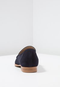 Mocassino in suede blu navy con tacco in legno basso, visto da dietro su una superficie bianca e uno sfondo grigio chiaro.