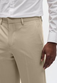 Pantalones beige a medida hechos de una tela suave, con frente plano, bolsillos laterales y un diseño de corte ajustado. Camisa blanca parcialmente visible en la parte superior.