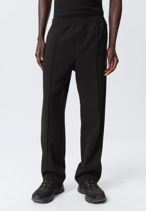 Svart treningsbukser med elastisk midje, med en løs passform og synlige sømmer. Matchet med svarte joggesko.