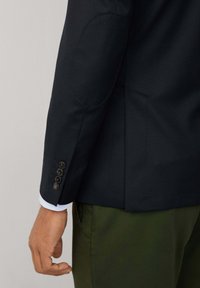 Blazer marine avec des coudières, détails de poignets à bouton et coupe ajustée. Porté sur une chemise bleu clair et associé à un pantalon vert olive.