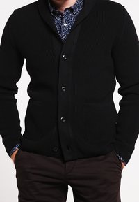 Schwarze, gerippte Strickjacke mit Schalkragen, Knopfverschluss, zwei vorderen Taschen und strukturiertem Stoff. Darüber wird ein blaues gemustertes Hemd getragen.