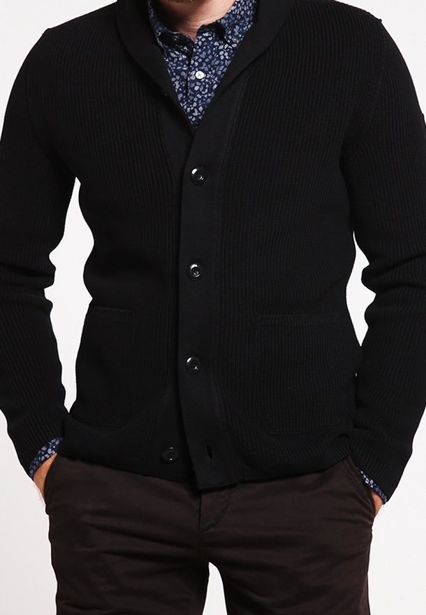 Schwarze, gerippte Strickjacke mit Schalkragen, Knopfverschluss, zwei vorderen Taschen und strukturiertem Stoff. Darüber wird ein blaues gemustertes Hemd getragen.