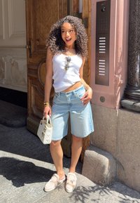 Top de malha branco com nervuras, calções de ganga azul claro, bolsa de mão creme, sapatos slip-on bege com detalhes em pelúcia. De pé contra uma porta de madeira.